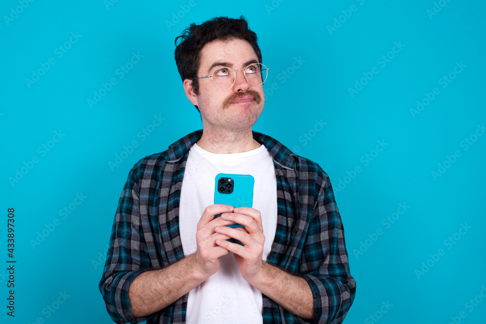 Portrait of young Caucasian man with moustache wearing plaid shirt against blue wall with dreamy look, thinking while holding smartphone. Tries to write up a message. Portrait of young Caucasian man with moustache wearing plaid shirt against blue wall with dreamy look, thinking while holding smartphone. Tries to write up a message.