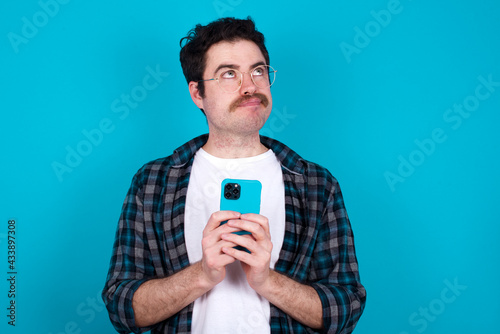 Portrait of young Caucasian man with moustache wearing plaid shirt against blue wall with dreamy look, thinking while holding smartphone. Tries to write up a message. © Jihan