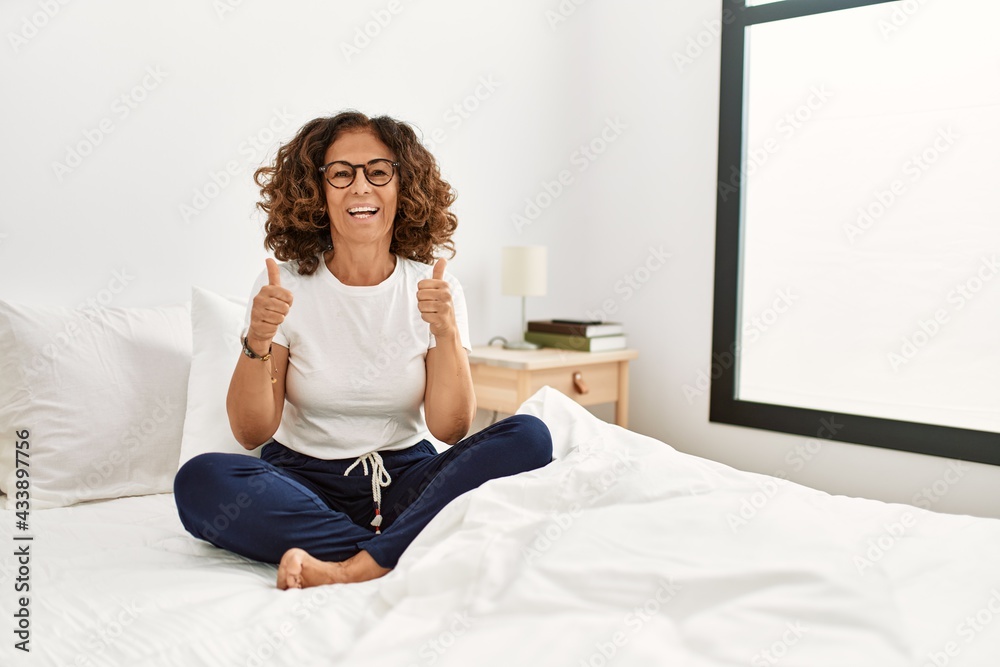 Middle age hispanic woman sitting on the bed at home success sign doing positive gesture with hand, thumbs up smiling and happy. cheerful expression and winner gesture.