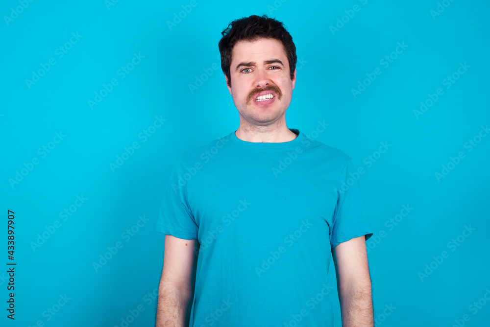 Portrait of dissatisfied young handsome Caucasian man with moustache wearing blue t-shirt smirks face, purses lips and looks with annoyance at camera, discontent hearing something unpleasant Portrait of dissatisfied young handsome Caucasian man with moustache wearing blue t-shirt smirks face, purses lips and looks with annoyance at camera, discontent hearing something unpleasant