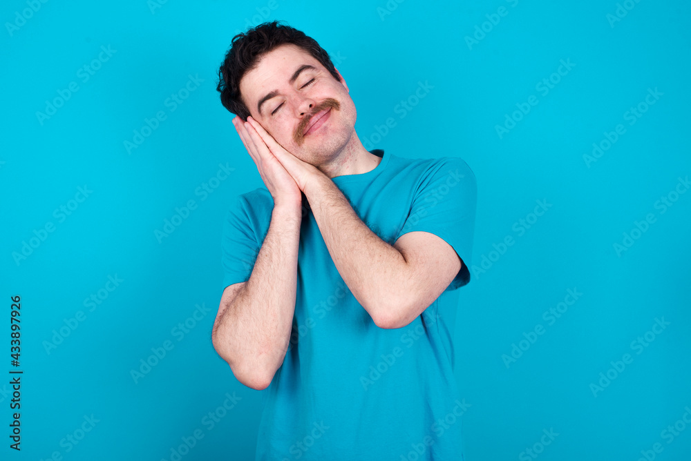 young handsome Caucasian man with moustache wearing blue t-shirt against blue background leans on pressed palms closes eyes and has pleasant smile dreams about something young handsome Caucasian man with moustache wearing blue t-shirt against blue background leans on pressed palms closes eyes and has pleasant smile dreams about something