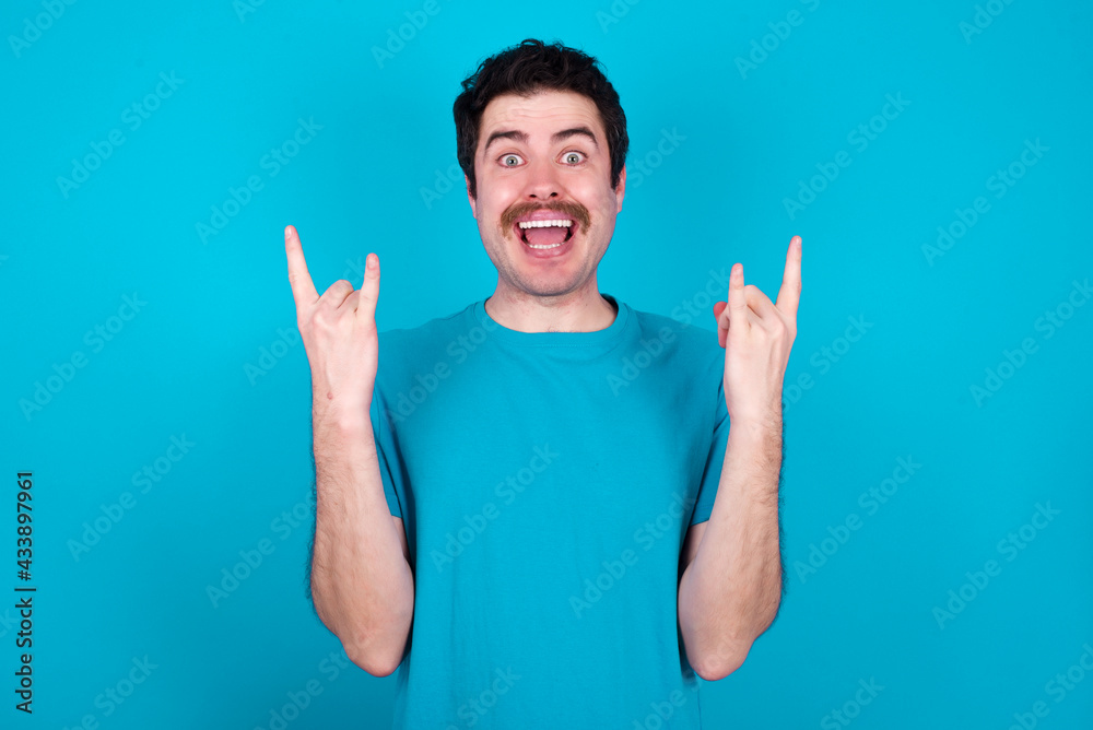 young handsome Caucasian man with moustache wearing blue t-shirt against blue background makes rock n roll sign looks self confident and cheerful enjoys cool music at party. Body language concept. young handsome Caucasian man with moustache wearing blue t-shirt against blue background makes rock n roll sign looks self confident and cheerful enjoys cool music at party. Body language concept.