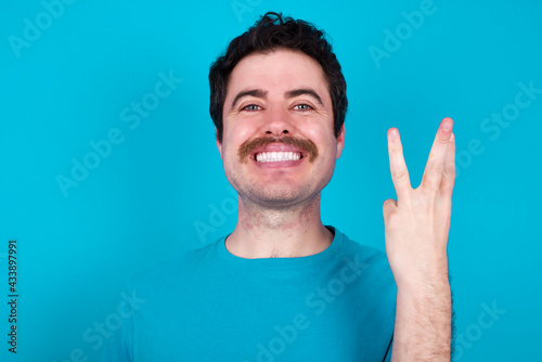 young handsome Caucasian man with moustache wearing blue t-shirt against blue background smiling and looking friendly, showing number three or third with hand forward, counting down