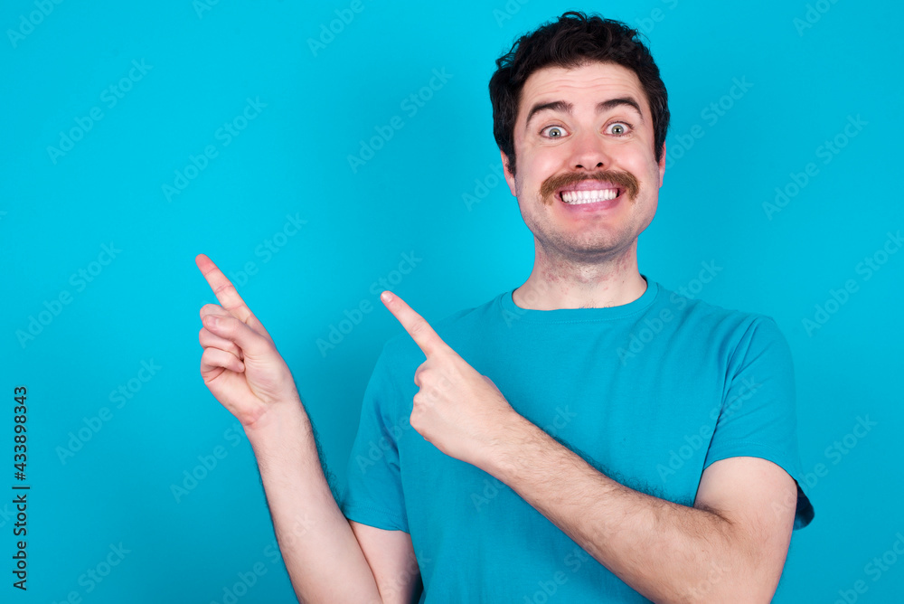 young handsome Caucasian man with moustache wearing blue t-shirt against blue background points at copy space indicates for advertising gives right direction young handsome Caucasian man with moustache wearing blue t-shirt against blue background points at copy space indicates for advertising gives right direction
