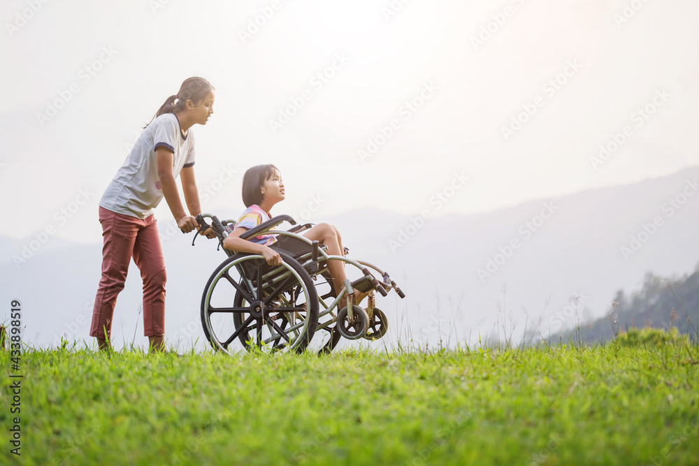 Obraz premium Disabled handicapped child sitting in wheelchair and care helper walking on mountain meadow park in sunny day.. International Disability Day.