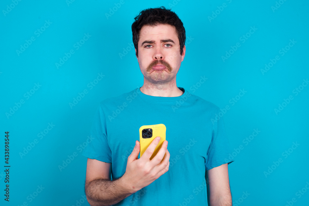 Upset dissatisfied young handsome Caucasian man with moustache wearing blue t-shirt against blue wall uses mobile software application and surrfs information in internet, holds modern mobile hand Upset dissatisfied young handsome Caucasian man with moustache wearing blue t-shirt against blue wall uses mobile software application and surrfs information in internet, holds modern mobile hand