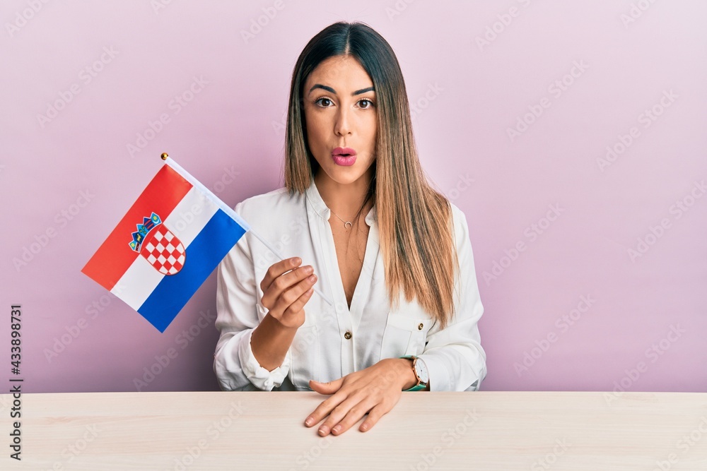 Young hispanic woman holding croatia flag sitting on the table scared and amazed with open mouth for surprise, disbelief face Young hispanic woman holding croatia flag sitting on the table scared and amazed with open mouth for surprise, disbelief face