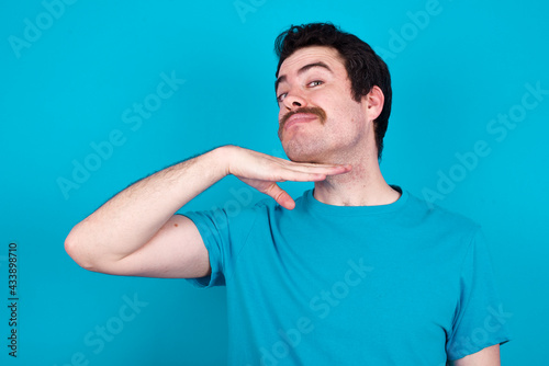 young handsome Caucasian man with moustache wearing blue t-shirt against blue background cutting throat with hand as knife, threaten aggression with furious violence.