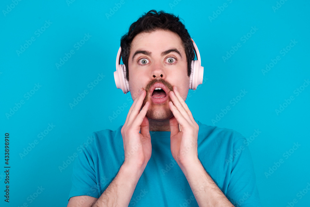 Shocked young handsome Caucasian man with moustache wearing blue t-shirt against blue wall stares fearful at camera keeps mouth widely opened wears wireless stereo headphones on ears Shocked young handsome Caucasian man with moustache wearing blue t-shirt against blue wall stares fearful at camera keeps mouth widely opened wears wireless stereo headphones on ears