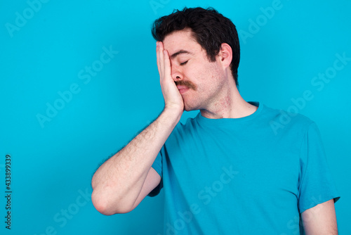 young handsome Caucasian man with moustache wearing blue t-shirt against blue background with sad expression covering face with hands while crying. Depression concept.