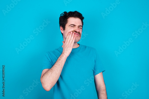 young handsome Caucasian man with moustache wearing blue t-shirt against blue background with toothache