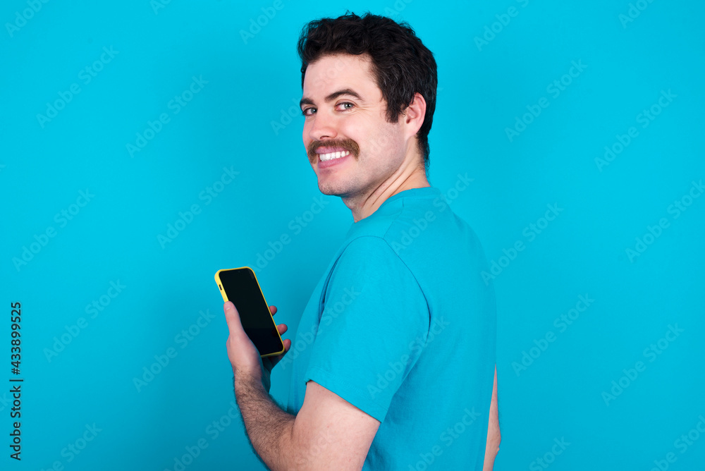 Rear view photo portrait of young handsome Caucasian man with moustache wearing blue t-shirt against blue background using smartphone smiling Rear view photo portrait of young handsome Caucasian man with moustache wearing blue t-shirt against blue background using smartphone smiling