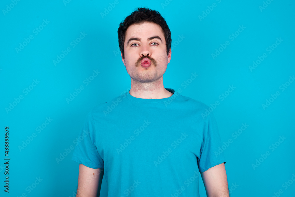 Portrait of lovely funny young handsome Caucasian man with moustache wearing orange t-shirt against blue background sending air kiss
