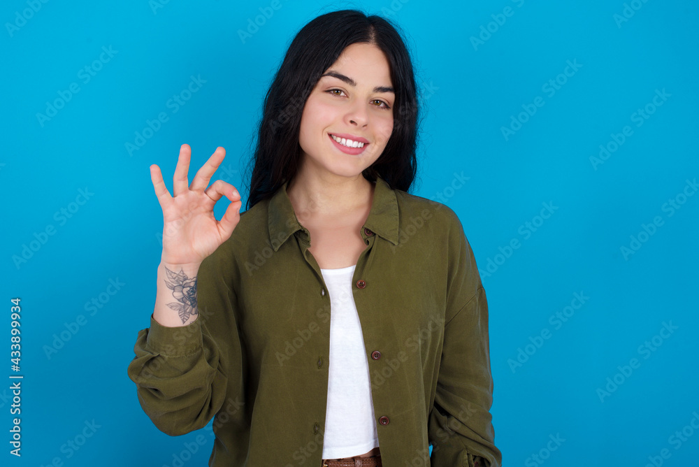 Glad attractive young beautiful tattooed girl standing against blue wall shows ok sign with hand as expresses approval, has cheerful expression, being optimistic. Glad attractive young beautiful tattooed girl standing against blue wall shows ok sign with hand as expresses approval, has cheerful expression, being optimistic.