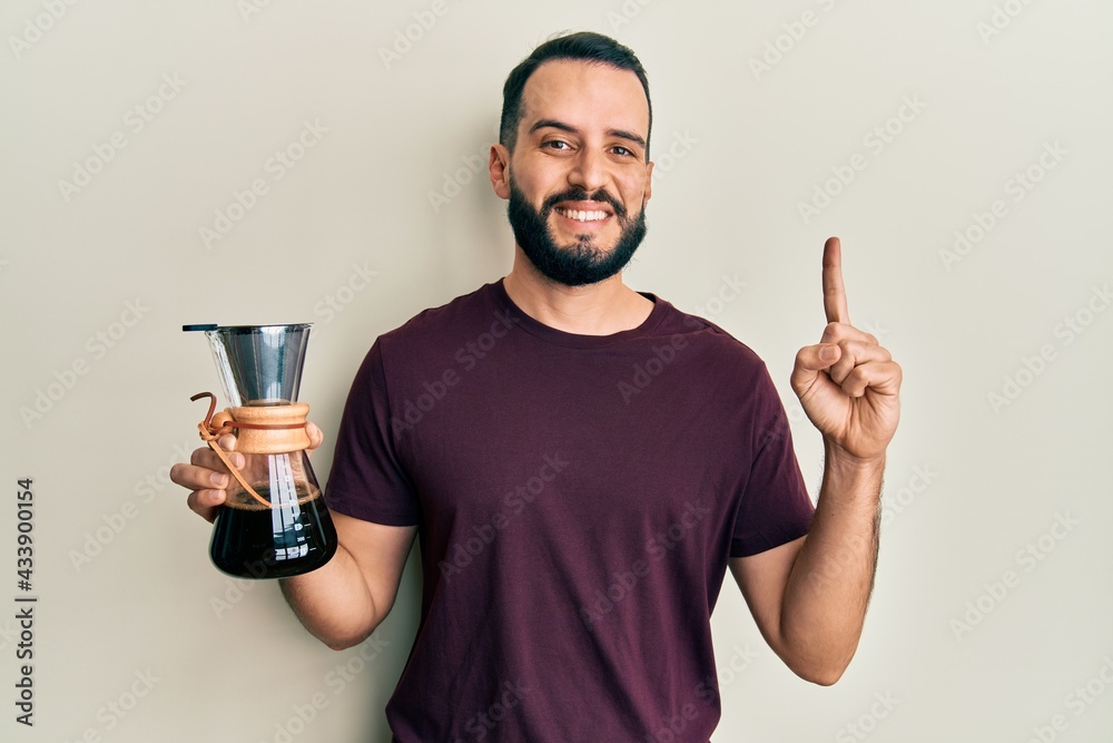 Young man with beard holding coffee maker with filter smiling with an idea or question pointing finger with happy face, number one Young man with beard holding coffee maker with filter smiling with an idea or question pointing finger with happy face, number one