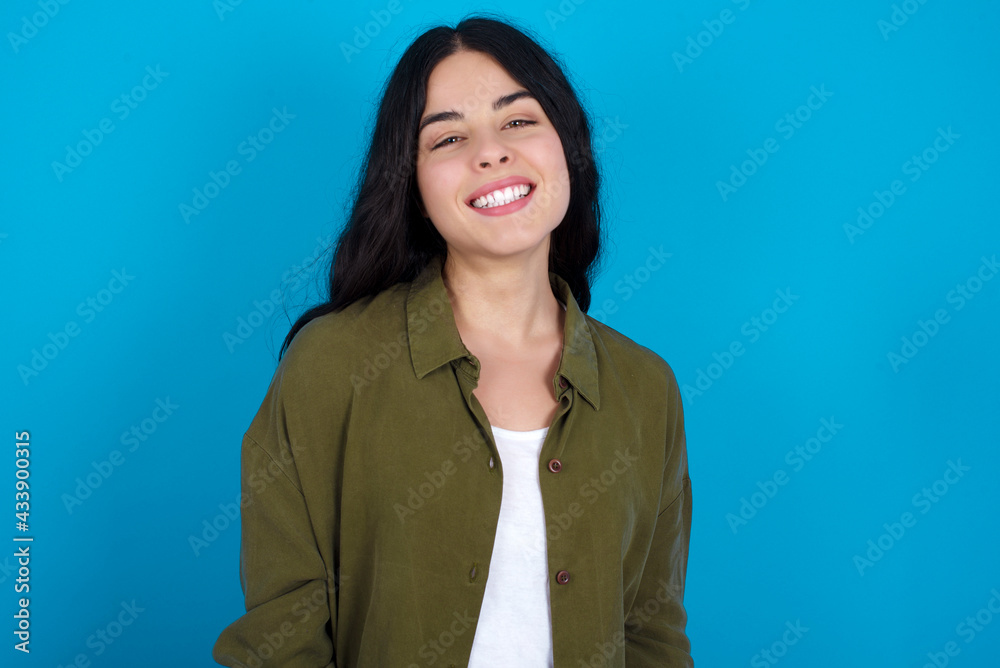 young beautiful tattooed girl standing against blue wall with broad smile, shows white teeth, feeling confident rejoices having day off. young beautiful tattooed girl standing against blue wall with broad smile, shows white teeth, feeling confident rejoices having day off.