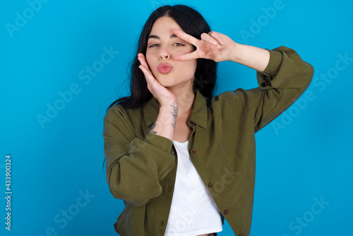 young beautiful tattooed girl standing against blue wall making v-sign near eyes. Leisure lifestyle people person celebrate flirt coquettish concept.