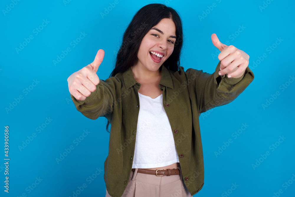 young beautiful tattooed girl standing against blue wall making positive gesture with thumbs up smiling and happy for success. Looking at the camera, winner gesture. young beautiful tattooed girl standing against blue wall making positive gesture with thumbs up smiling and happy for success. Looking at the camera, winner gesture.
