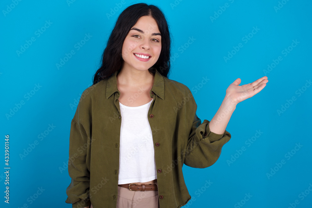 Positive glad young beautiful tattooed girl standing against blue background says: wow how exciting it is, has amazed expression, shows something on blank space with open hand. Advertisement concept.
