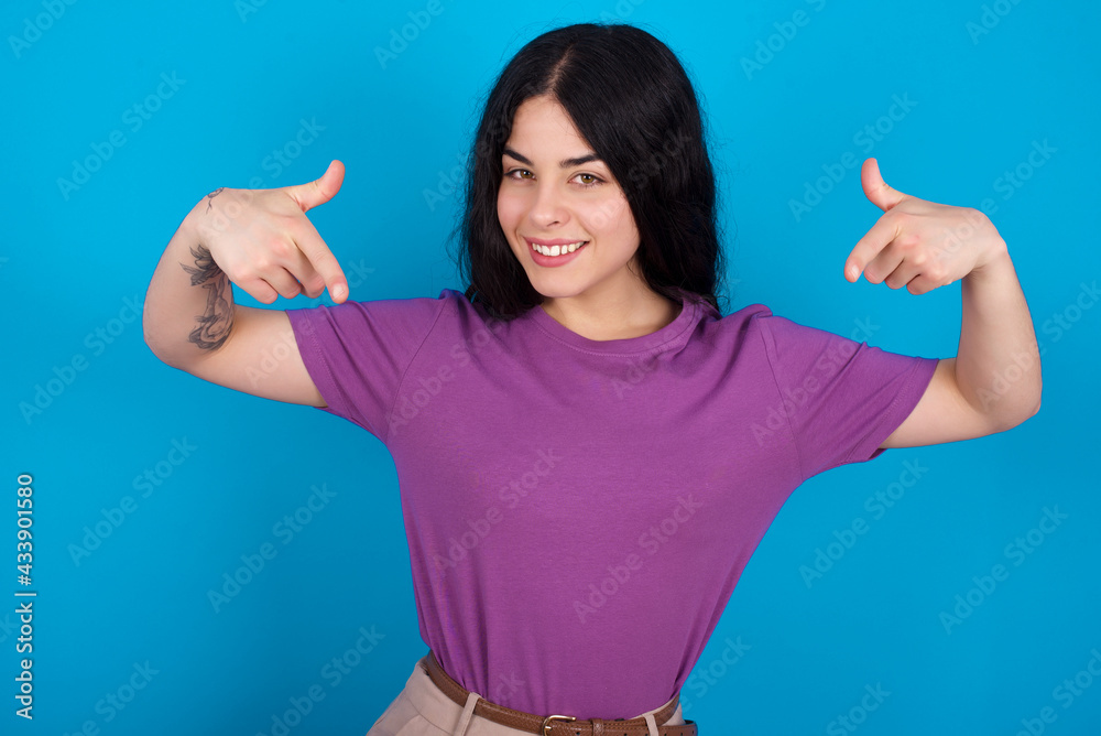 Pick me! Confident, self-assured and charismatic young beautiful tattooed girl wearing purple t-shirt standing against blue wall promoting oneself as wanting role smiling broadly and pointing at body. Pick me! Confident, self-assured and charismatic young beautiful tattooed girl wearing purple t-shirt standing against blue wall promoting oneself as wanting role smiling broadly and pointing at body.