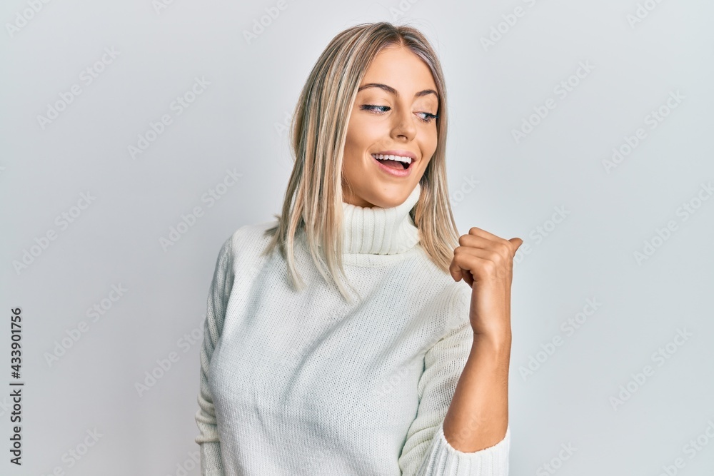 Beautiful blonde woman wearing casual turtleneck sweater pointing thumb up to the side smiling happy with open mouth Beautiful blonde woman wearing casual turtleneck sweater pointing thumb up to the side smiling happy with open mouth