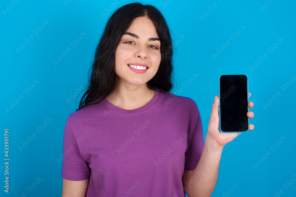 Smiling young beautiful tattooed girl wearing purple t-shirt standing against blue background showing empty phone screen. Advertisement and communication concept. Smiling young beautiful tattooed girl wearing purple t-shirt standing against blue background showing empty phone screen. Advertisement and communication concept.