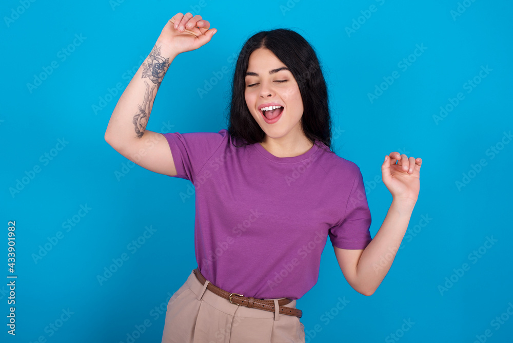 Photo of upbeat young beautiful tattooed girl wearing purple t-shirt standing against blue has fun and dances carefree wear being in perfect mood makes movements. Spends free time on disco party Photo of upbeat young beautiful tattooed girl wearing purple t-shirt standing against blue has fun and dances carefree wear being in perfect mood makes movements. Spends free time on disco party