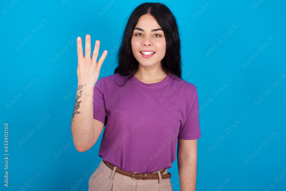 young beautiful tattooed girl wearing purple t-shirt standing against blue background smiling and looking friendly, showing number four or fourth with hand forward, counting down young beautiful tattooed girl wearing purple t-shirt standing against blue background smiling and looking friendly, showing number four or fourth with hand forward, counting down
