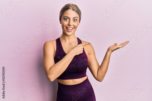 Beautiful blonde woman wearing sportswear over pink background amazed and smiling to the camera while presenting with hand and pointing with finger. © Krakenimages.com