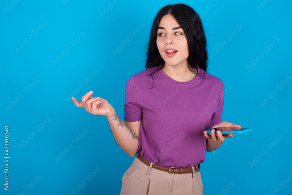 Happy pleased young beautiful tattooed girl wearing blue t-shirt standing against blue background raises palm and holds cellphone uses high speed internet for text messaging or video calls Happy pleased young beautiful tattooed girl wearing blue t-shirt standing against blue background raises palm and holds cellphone uses high speed internet for text messaging or video calls