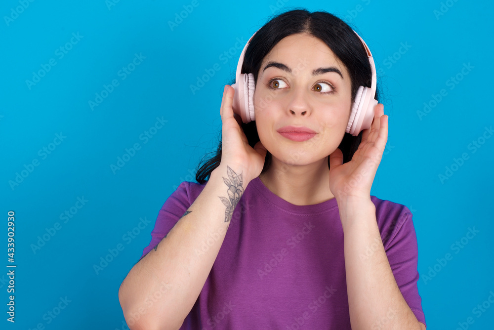 young beautiful tattooed girl wearing blue t-shirt standing against blue background wears stereo headphones listens music concentrated aside. People hobby lifestyle concept young beautiful tattooed girl wearing blue t-shirt standing against blue background wears stereo headphones listens music concentrated aside. People hobby lifestyle concept