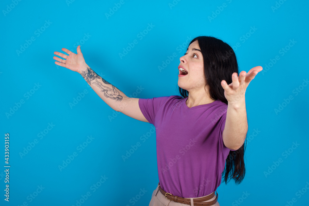 Funny astonished young beautiful tattooed girl wearing purple t-shirt standing against blue background look empty space with arms opened ready to catch something. Funny astonished young beautiful tattooed girl wearing purple t-shirt standing against blue background look empty space with arms opened ready to catch something.