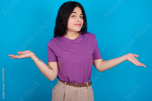 Careless attractive young beautiful tattooed girl wearing purple t-shirt standing against blue background shrugging shoulders, oops.