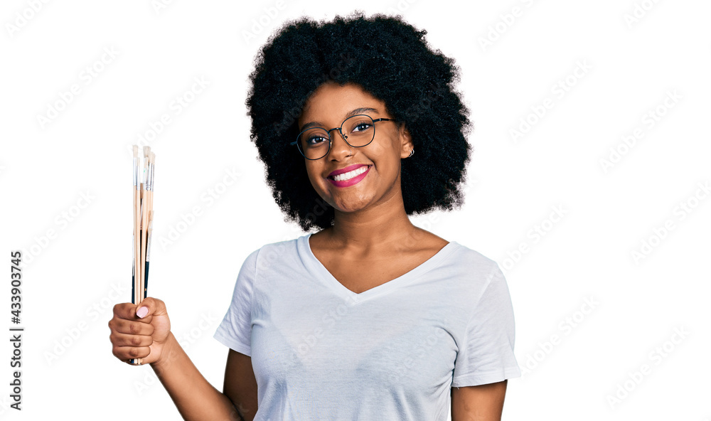 Young african american woman holding paintbrushes looking positive and happy standing and smiling with a confident smile showing teeth Young african american woman holding paintbrushes looking positive and happy standing and smiling with a confident smile showing teeth