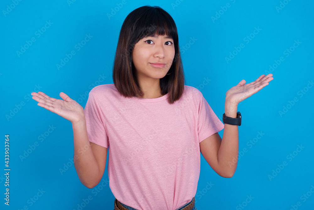 Maybe yes or no. Doubtful young beautiful asian woman wearing pink t-shirt against blue wall shrugs shoulders in bewilderment, tries to make decision puzzled what he wants. Hesitation and uncertainty. Maybe yes or no. Doubtful young beautiful asian woman wearing pink t-shirt against blue wall shrugs shoulders in bewilderment, tries to make decision puzzled what he wants. Hesitation and uncertainty.