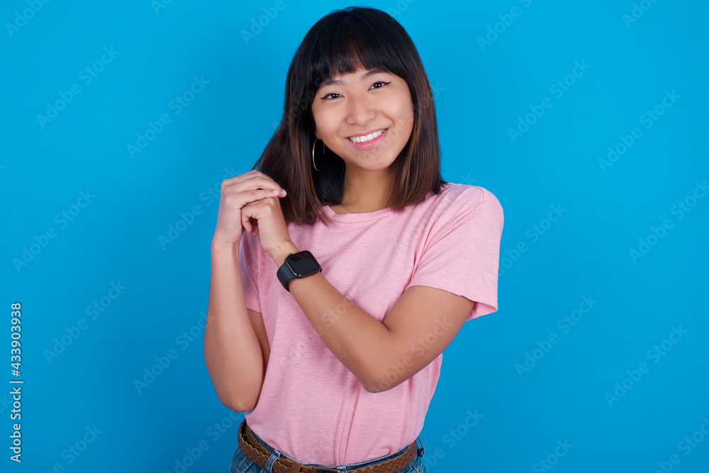 Dreamy young beautiful asian woman wearing pink t-shirt against blue wall with pleasant expression, closes eyes, keeps hands crossed near face, thinks about something pleasant Dreamy young beautiful asian woman wearing pink t-shirt against blue wall with pleasant expression, closes eyes, keeps hands crossed near face, thinks about something pleasant