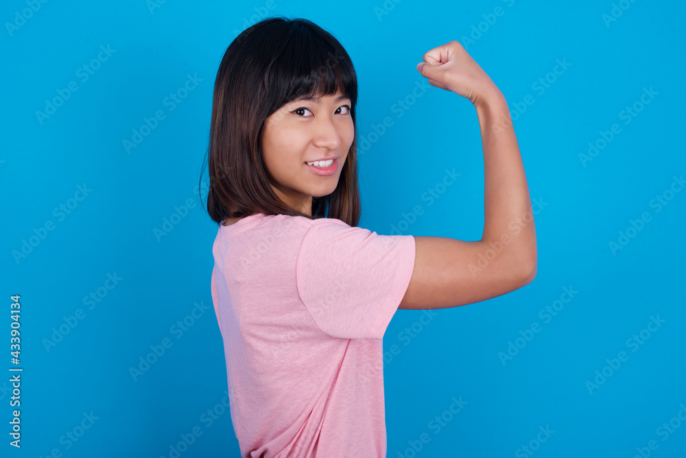young beautiful asian woman wearing pink t-shirt against blue wall, showing muscles after workout. Health and strength concept. young beautiful asian woman wearing pink t-shirt against blue wall, showing muscles after workout. Health and strength concept.