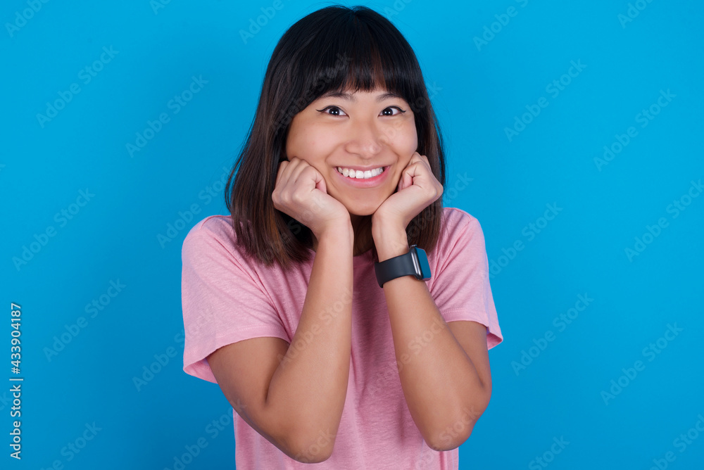 Dreamy young beautiful asian woman wearing pink t-shirt against blue wall keeps hands pressed together under chin, looks with happy expression, has toothy smile. Dreamy young beautiful asian woman wearing pink t-shirt against blue wall keeps hands pressed together under chin, looks with happy expression, has toothy smile.
