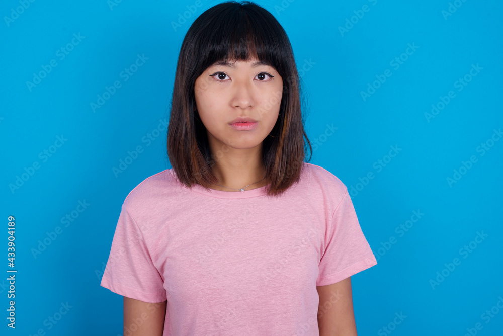 Joyful young beautiful asian woman wearing pink t-shirt against blue wall looking to the camera, thinking about something. Both arms down, neutral facial expression. Joyful young beautiful asian woman wearing pink t-shirt against blue wall looking to the camera, thinking about something. Both arms down, neutral facial expression.