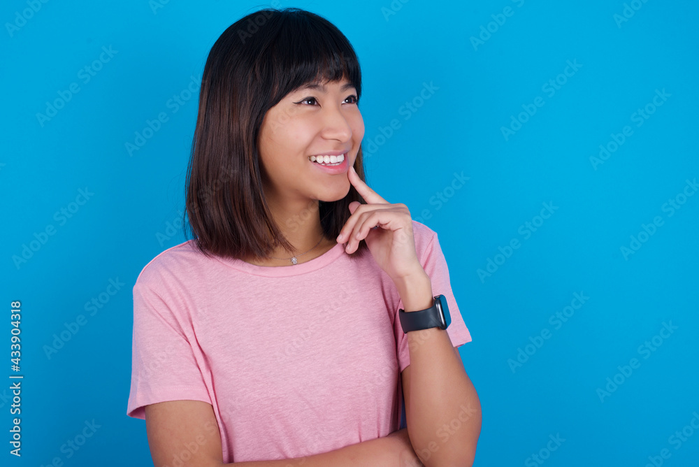 young beautiful asian woman wearing pink t-shirt against blue wall with thoughtful expression, looks to the camera, keeps hand near face, bitting a finger thinks about something pleasant. young beautiful asian woman wearing pink t-shirt against blue wall with thoughtful expression, looks to the camera, keeps hand near face, bitting a finger thinks about something pleasant.