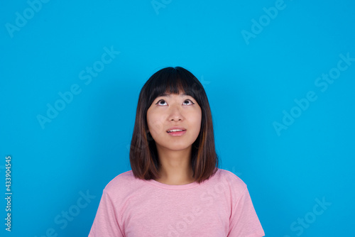 Fotografija young beautiful asian woman wearing pink t-shirt against blue wall looking up as he sees something strange