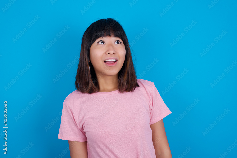Surprised young beautiful asian woman wearing purple t-shirt against blue wall, shrugs shoulders, looking sideways, being happy and excited. Sudden reactions concept. Surprised young beautiful asian woman wearing purple t-shirt against blue wall, shrugs shoulders, looking sideways, being happy and excited. Sudden reactions concept.