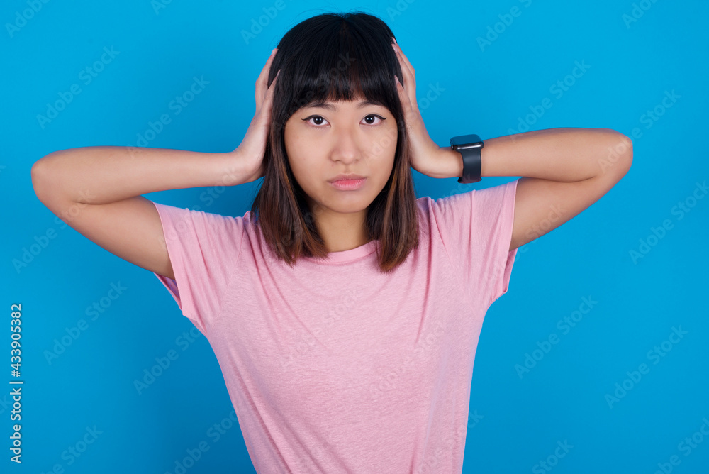 Frustrated young beautiful asian woman wearing purple t-shirt against blue wall plugging ears with hands does not wanting to listen hard rock, noise or loud music. Frustrated young beautiful asian woman wearing purple t-shirt against blue wall plugging ears with hands does not wanting to listen hard rock, noise or loud music.