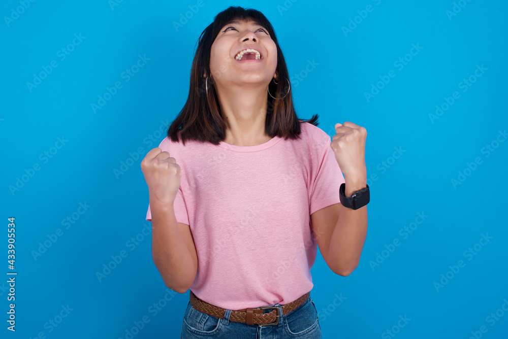 Young beautiful asian girl wearing pink t-shirt against blue background looks with excitement up, keeps hands raised, notices something unexpected. Young beautiful asian girl wearing pink t-shirt against blue background looks with excitement up, keeps hands raised, notices something unexpected.