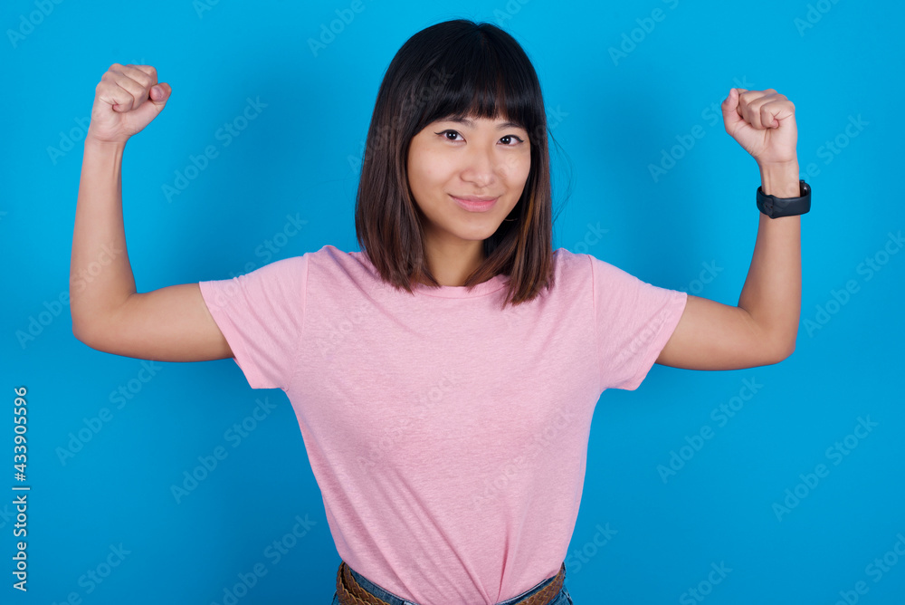 Waist up shot of Young beautiful asian girl wearing pink t-shirt raises arms to show muscles feels confident in victory, looks strong and independent, smiles positively at camera. Sport concept. Waist up shot of Young beautiful asian girl wearing pink t-shirt raises arms to show muscles feels confident in victory, looks strong and independent, smiles positively at camera. Sport concept.