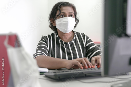 Asian businesswoman wearing face mask typing on keyboard inside office.