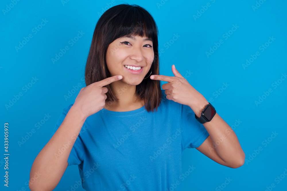 Strong healthy straight white teeth. Close up portrait of happy Young beautiful asian girl wearing blue t-shirt against blue background with beaming smile pointing on perfect clear white teeth. Strong healthy straight white teeth. Close up portrait of happy Young beautiful asian girl wearing blue t-shirt against blue background with beaming smile pointing on perfect clear white teeth.