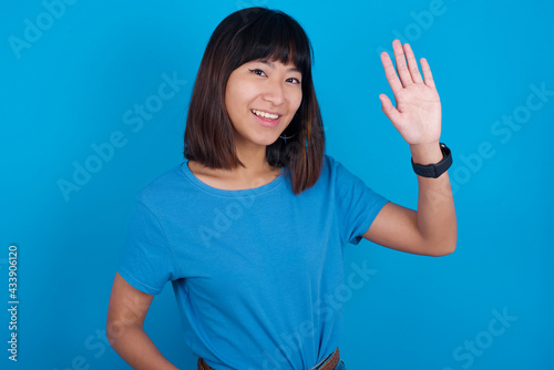 Young beautiful asian girl wearing blue t-shirt against blue background waiving saying hello or goodbye happy and smiling, friendly welcome gesture.