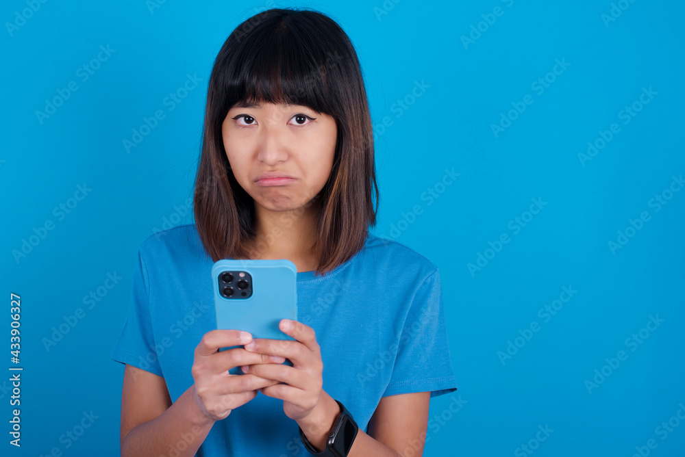Portrait of a confused Young beautiful asian girl wearing purple t-shirt against blue background holding mobile phone and shrugging shoulders and frowning face.