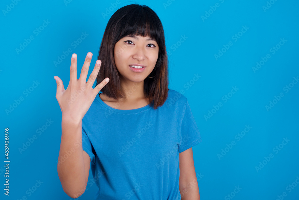 young beautiful asian girl wearing blue t-shirt against blue background smiling and looking friendly, showing number five or fifth with hand forward, counting down young beautiful asian girl wearing blue t-shirt against blue background smiling and looking friendly, showing number five or fifth with hand forward, counting down